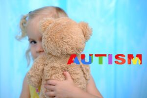 תמונת נושא של המאמר על תביעת ביטוח סיעודי אוטיזם המציגה ילדה קטנה עם שיער בלונדיני מחזיקה דובון פרווה המסתיר את פניה ברקע כחול עם הטקסט הצבעוני AUTISM לצד הדובה.